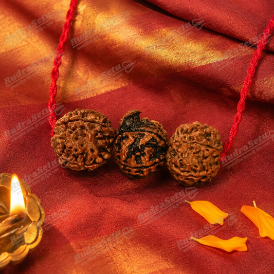 Three rudraksha beads on a red cord on deep red silk, beside a lit brass diya and marigold petals